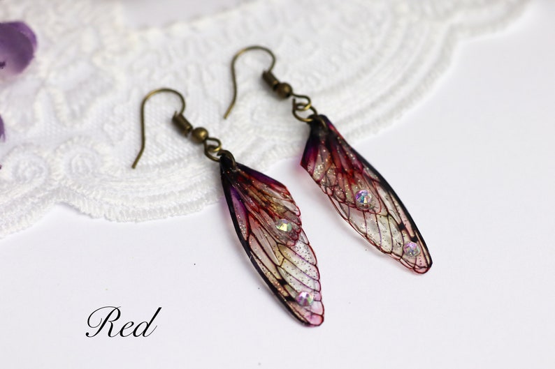 May include: A pair of earrings featuring translucent wings with a red, purple, and black color scheme. The wings have a delicate, intricate design and are accented with small, iridescent rhinestones.