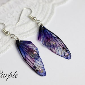May include: A pair of dangle earrings featuring iridescent purple and blue wings with silver glitter and rhinestones. The earrings are labeled "Purple".