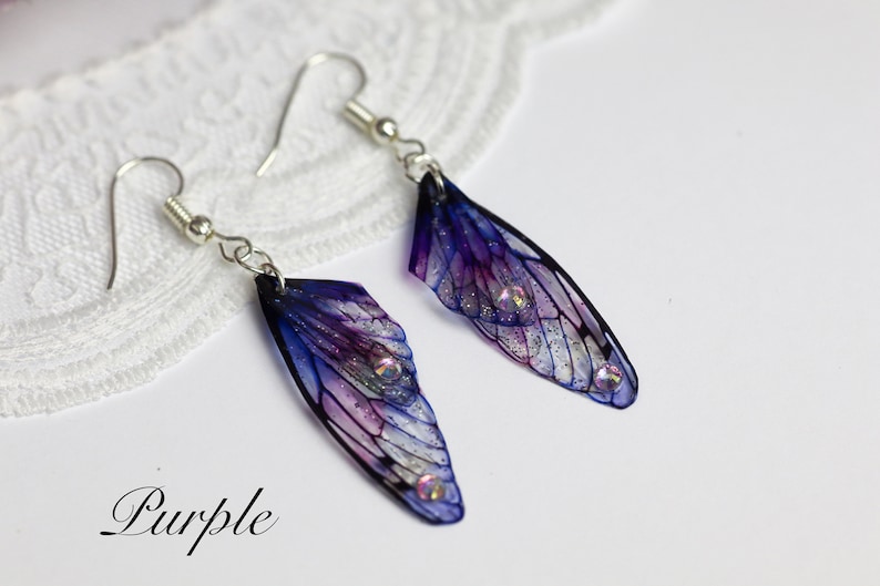 May include: A pair of dangle earrings featuring iridescent purple and blue wings with silver glitter and rhinestones. The earrings are labeled "Purple".