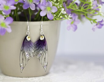 May include: A pair of purple and silver fairy wing earrings with clear crystal accents. The earrings are hanging from a silver hook.