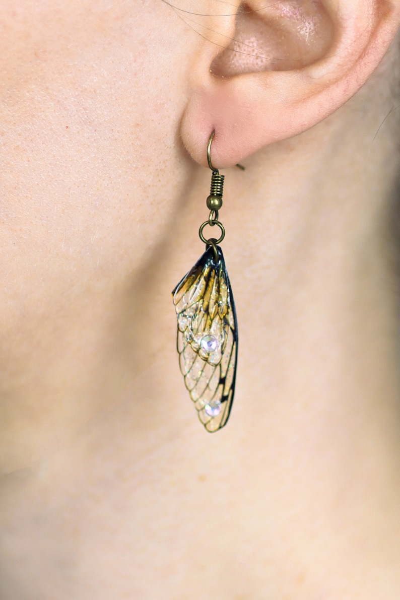 May include: A pair of earrings featuring a delicate, translucent insect wing design with a bronze-colored metal hook. The wings are accented with small, clear rhinestones.