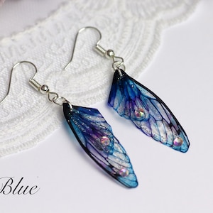 May include: A pair of blue and purple fairy wing earrings with silver hooks. The wings have a translucent, iridescent finish and are decorated with glitter and rhinestones.