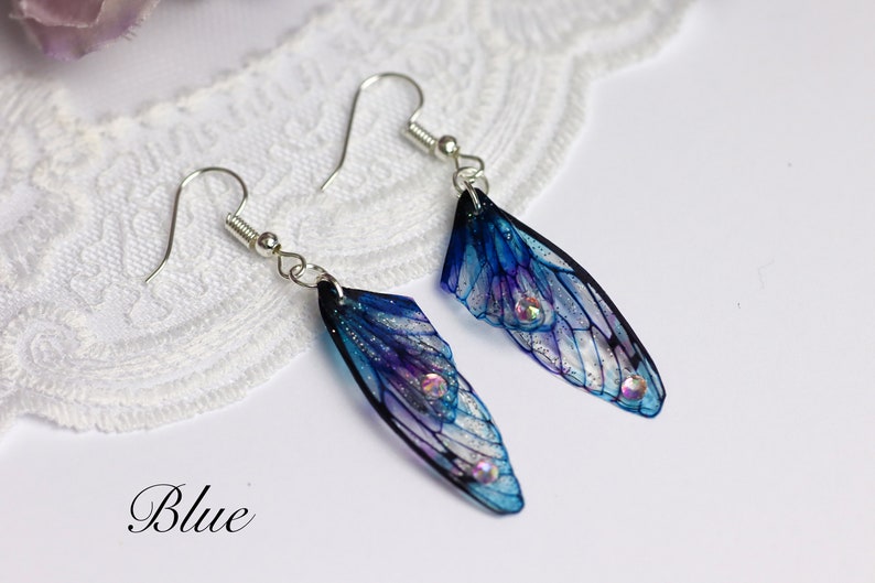 May include: A pair of blue and purple fairy wing earrings with silver hooks. The wings have a translucent, iridescent finish and are decorated with glitter and rhinestones.