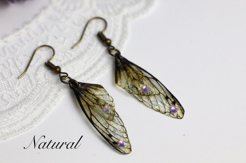 May include: A pair of earrings featuring delicate, translucent insect wings with iridescent accents. The wings are a light brown color with a subtle shimmer. The earrings are suspended from bronze-colored hooks.