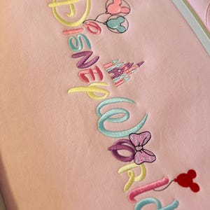 May include: Pink fabric embroidered with the words "Disney World" in various pastel colors. The design includes Mickey Mouse balloons, a castle, and a Minnie Mouse bow. The embroidery is detailed and colorful.