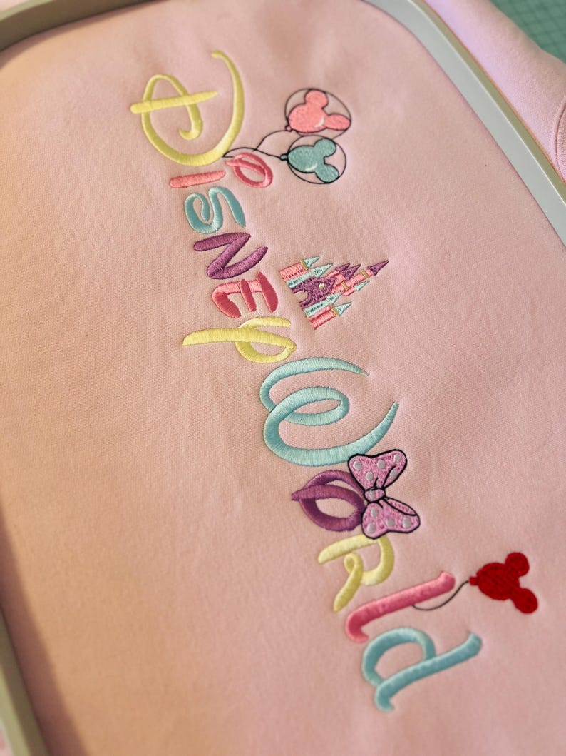 May include: Pink fabric embroidered with the words "Disney World" in various pastel colors. The design includes Mickey Mouse balloons, a castle, and a Minnie Mouse bow. The embroidery is detailed and colorful.