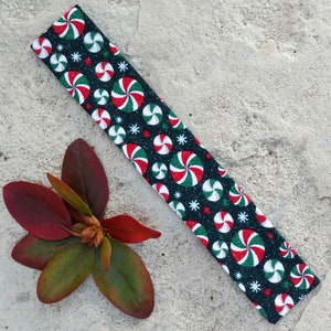 May include: A rectangular fabric headband featuring a black background with a repeating pattern of red, white, and green peppermint candy swirls and small white starburst shapes. The fabric has a subtle glittery finish throughout the design.