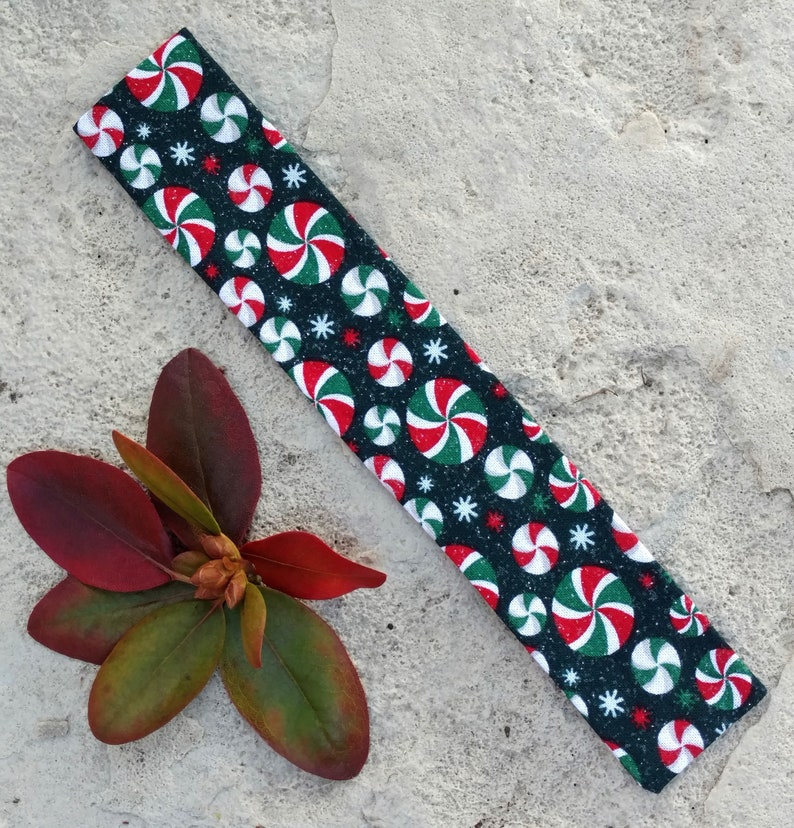 May include: A rectangular fabric headband featuring a black background with a repeating pattern of red, white, and green peppermint candy swirls and small white starburst shapes. The fabric has a subtle glittery finish throughout the design.