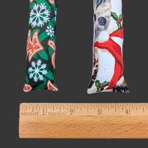 May include: Two rectangular fabric cat toys positioned above a wooden Westcott ruler. The left toy features a green background with white snowflakes and orange gingerbread shapes. The right toy displays a cat wearing a red scarf and holly leaves.