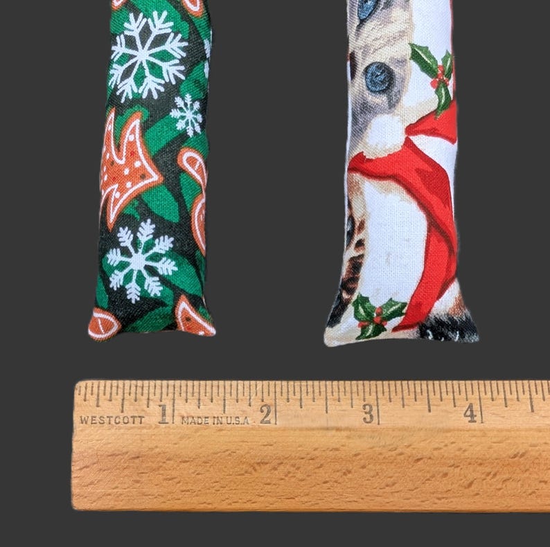 May include: Two rectangular fabric cat toys positioned above a wooden Westcott ruler. The left toy features a green background with white snowflakes and orange gingerbread shapes. The right toy displays a cat wearing a red scarf and holly leaves.