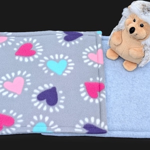 May include: A gray fleece pet bed with a pink, blue, and purple heart pattern. A plush hedgehog toy rests on the bed. The bed is rectangular and has a solid light blue fleece side.