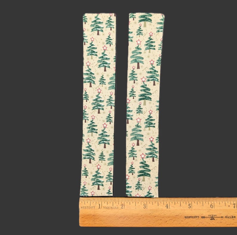 May include: Two rectangular fabric strips featuring a repeating pattern of green Christmas trees with red star toppers on a cream background. A wooden ruler is positioned at the base, showing each strip is approximately 6.5 inches in length.