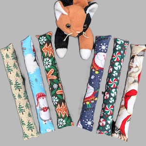 May include: A plush calico cat toy positioned above six long, rectangular fabric cat kicker toys. The kickers feature various holiday patterns including green pine trees, snowmen, gingerbread men, Santa Claus, peppermint candies, and cats.