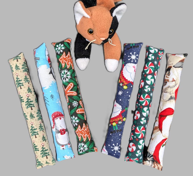 May include: A plush calico cat toy positioned above six long, rectangular fabric cat kicker toys. The kickers feature various holiday patterns including green pine trees, snowmen, gingerbread men, Santa Claus, peppermint candies, and cats.