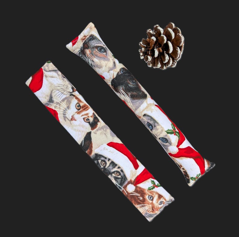 May include: Two long, rectangular fabric strips featuring a repeating print of cats wearing red Santa hats, accompanied by a single brown pinecone with white-tipped scales. The fabric pattern includes various cat faces and festive holly accents.
