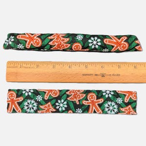 May include: Two rectangular fabric strips featuring a black background with a repeating pattern of orange gingerbread people, white snowflakes, and orange Christmas trees. A wooden Westcott ruler is placed between them, showing a length of 9 inches.
