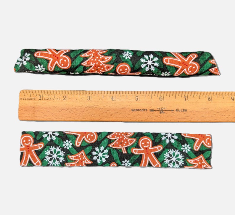 May include: Two rectangular fabric strips featuring a black background with a repeating pattern of orange gingerbread people, white snowflakes, and orange Christmas trees. A wooden Westcott ruler is placed between them, showing a length of 9 inches.