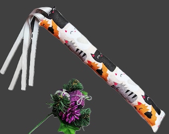 May include: A long, cylindrical fabric toy featuring a repeating pattern of cartoon cats in black, white, orange, and gray. Three white ribbons extend from one end. Beside it is a decorative cluster of green pine needles and a purple pinecone.