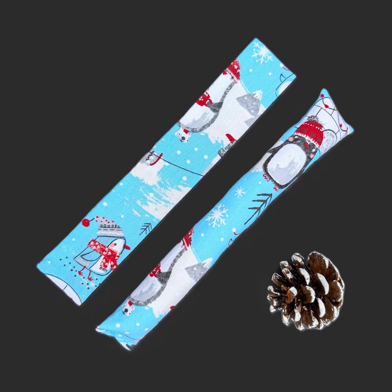 May include: Two long, rectangular fabric strips featuring a light blue background with a repeating pattern of penguins wearing red hats and scarves, white snowflakes, and mountains. A single brown pinecone is positioned near the fabric strips.