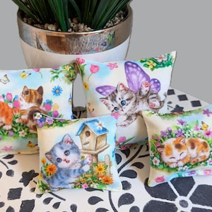 May include: Four small square pillows featuring colorful illustrations of kittens in garden settings. The designs include kittens with butterflies, a kitten near a birdhouse, and a sleeping kitten, all surrounded by vibrant flowers and greenery.
