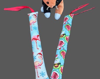 May include: A plush calico cat toy positioned above two long, rectangular fabric cat kicker toys. One kicker features a light blue background with pink flamingos wearing Santa hats and snowflakes. The other features avocados wearing Santa hats.