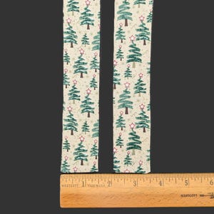 May include: Two strips of off-white fabric featuring a repeating pattern of green pine trees and red stars. A wooden ruler is positioned horizontally at the base, showing the fabric strips are approximately 2 inches wide each.