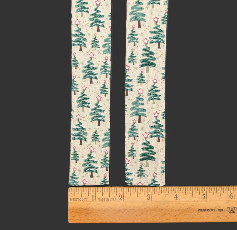 May include: Two strips of off-white fabric featuring a repeating pattern of green pine trees and red stars. A wooden ruler is positioned horizontally at the base, showing the fabric strips are approximately 2 inches wide each.