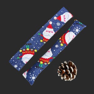 May include: Two long, rectangular fabric strips featuring a dark blue background with a repeating pattern of Santa Claus figures, white snowflakes, and the text Ho Ho Ho. A single brown pinecone is positioned near the bottom right corner.