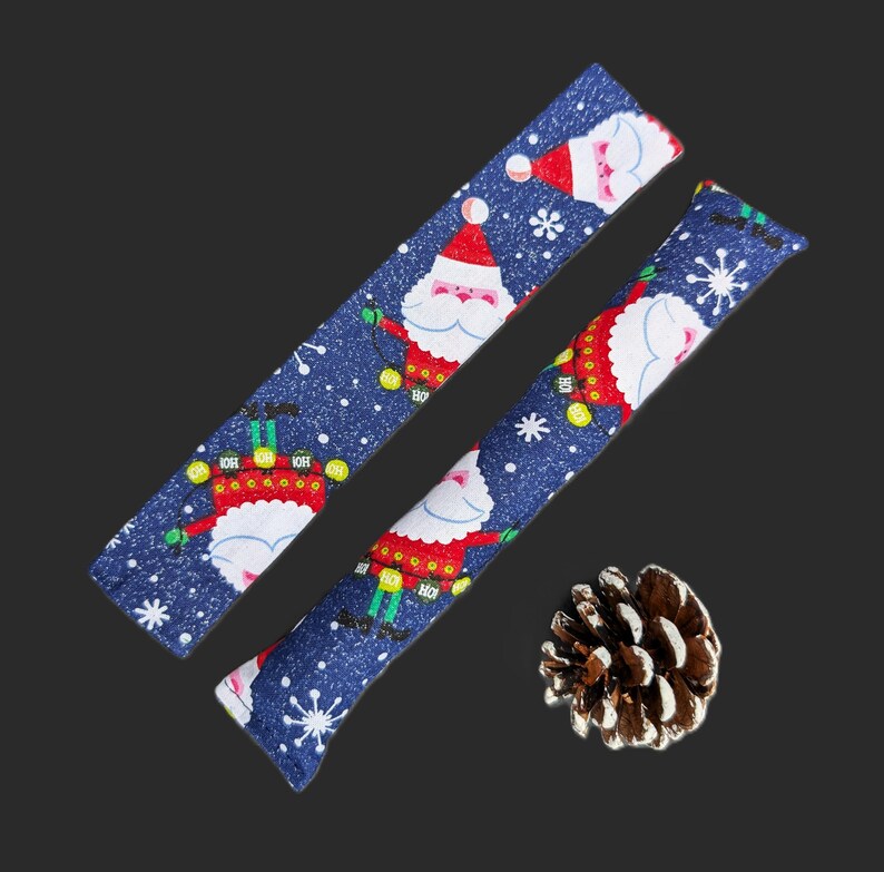 May include: Two long, rectangular fabric strips featuring a dark blue background with a repeating pattern of Santa Claus figures, white snowflakes, and the text Ho Ho Ho. A single brown pinecone is positioned near the bottom right corner.