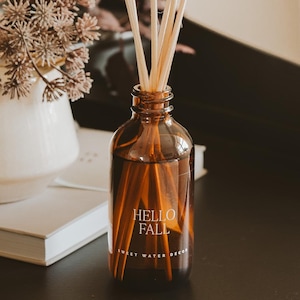 May include: A brown glass diffuser bottle with reed sticks, labeled "Hello Fall Sweet Water Decor." The bottle sits on a dark surface next to a white vase with dried flowers and a stack of books. The scene evokes a cozy, autumnal atmosphere.