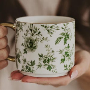 May include: White ceramic mug with a gold handle and rim, adorned with a green floral pattern. The mug is held by a person. The floral design features various green leaves and flowers, creating a botanical theme.