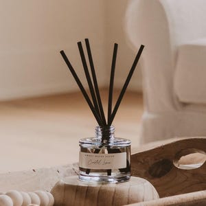 May include: A clear glass essential oil diffuser with black reeds, labeled "Sweet Water Decor, Coastal Linen." The diffuser sits on a wooden stand inside a wooden tray with decorative wooden beads. The tray is on a neutral-colored rug.