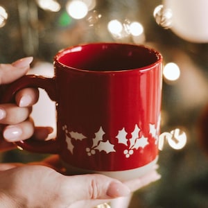 May include: A red ceramic mug with a white holly design is held in front of a Christmas tree. The mug has a handle and a light-colored base. The background features blurred Christmas lights and ornaments.
