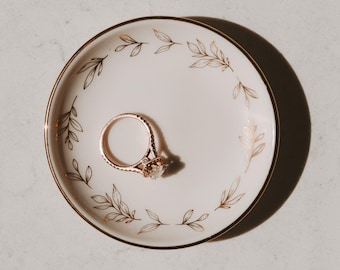 May include: A gold ring with a large, clear gemstone rests on a small, round, off-white dish. The dish has a gold rim and a delicate leaf pattern in gold around the edge. The ring is centered on the dish.