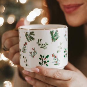 May include: A white ceramic mug with a gold rim, decorated with green sprigs and red berries. The mug is held by a person, with a blurred background of Christmas lights.