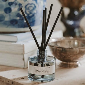May include: A clear glass diffuser bottle with black reeds and a label that reads "Shore Lily" sits on a wooden surface. A blue and white patterned vase and a stack of books are in the background, along with a decorative bowl.