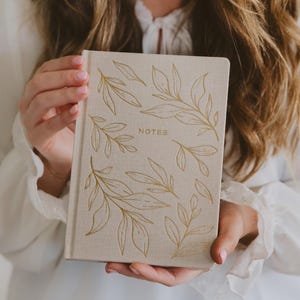 May include: A beige notebook with gold leaf designs and the word "NOTES" printed on the cover. The notebook is held by a person wearing a white blouse with ruffled sleeves. The design features several gold leaves.