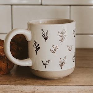 May include: A white ceramic mug with a beige base and a large handle. The mug is decorated with a pattern of black leaf illustrations. A muffin is visible to the left of the mug, on a wooden surface.