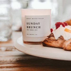 May include: A white candle with a label that reads "Sweet Water Decor Sunday Brunch Apple + Bourbon + Maple 6.5 oz | Made in USA | 11 oz". The candle is on a white plate with a pastry topped with whipped cream and raspberries.