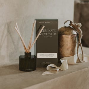 May include: A black glass reed diffuser with wooden reeds, a dark green rectangular box labeled ILLUMINATE EVERYDAY REED DIFFUSER SNOW COVERED PINES 3.5 FL OZ (104 ML), and a rustic bronze-colored metal bell with a rope handle and cream ribbon.