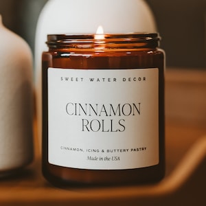 May include: A lit amber glass candle with a white label sits on a wooden surface between two white ceramic vases. The label reads 'Sweet Water Decor,' 'Cinnamon Rolls,' 'Cinnamon, Icing & Buttery Pastry,' and 'Made in the USA.' The candle and vases are home decor items with a minimalist aesthetic.