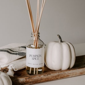 May include: A clear glass bottle with a white label that reads "Pumpkin Spice" and a wooden diffuser stick. The bottle is sitting on a wooden tray with two white pumpkins.
