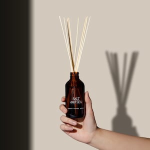 May include: A brown glass bottle diffuser with a white label that reads "SALT and SEA" and "SWEET WATER REED DIFFUSER". The bottle is filled with a clear liquid and has natural wooden sticks inserted into it.
