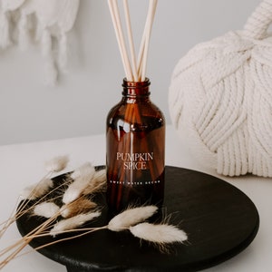 May include: An amber glass reed diffuser bottle with a label reading PUMPKIN SPICE SWEET WATER DECOR. The bottle contains several light-colored wooden reeds and is placed on a round black wooden tray alongside dried beige bunny tail grass stems.