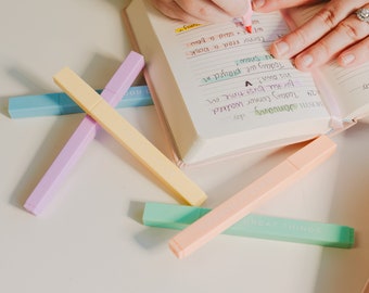 May include: A close-up of a hand writing in a pink journal with a pink highlighter.  There are five pastel colored highlighters on a white surface. The highlighters are labeled "GOOD LIFE" and "GREAT THINGS".