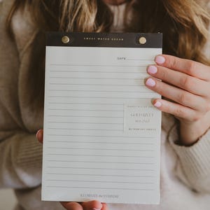 May include: A lined notepad with a black top and gold rivets. The notepad has the text "SWEET WATER DECOR" at the top and "GOLD RIVET notepad" on the bottom right. The notepad is held by a person.