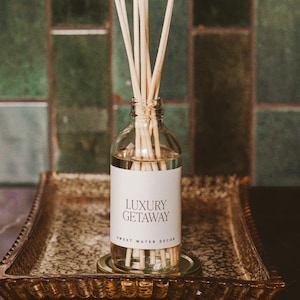 May include: A clear glass bottle with a white label that reads "Luxury Getaway" and "Sweet Water Reed Diffuser". The bottle is filled with a clear liquid and has wooden sticks inserted into it. The bottle is sitting on a gold-colored tray with a decorative design. The background is a green tiled wall.