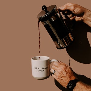 May include: A white ceramic mug with the text "Best Dad Ever" is being filled with coffee from a silver french press.