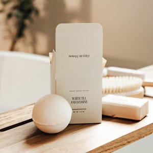 May include: A white bath bomb and its packaging are displayed on a wooden surface. The packaging reads "White Tea and Jasmine" and "Enjoy the Moment." The bath bomb is a sphere, and the box is open, revealing the product.