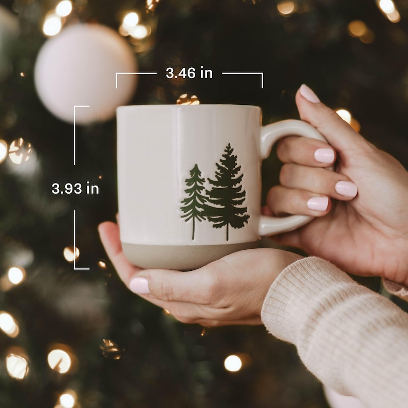 May include: A cream-colored ceramic mug featuring a dark green pine tree graphic on the front. The mug has a smooth upper finish and a matte base. Dimensions are 3.46 inches in width and 3.93 inches in height as indicated by the text overlay.